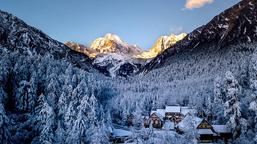 Snow-covered village nestled in a forest, with majestic sunlit mountains in the background and a clear blue sky above.