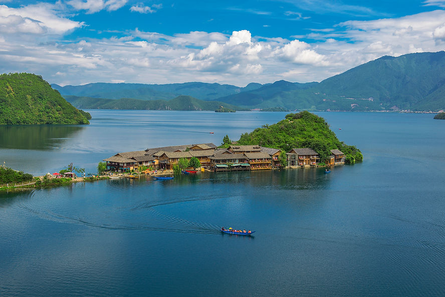 Peaceful lakeside village with wooden houses nestled on green hills. A boat with people glides on the calm water under a blue sky.