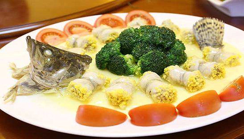 Fish head and tail arranged on a plate with broccoli, rolled fish, and tomato slices. Creamy yellow sauce adds a vibrant touch.