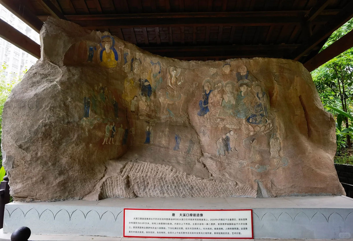 Ancient rock carving of colorful figures under a wooden shelter, with greenery in the background and a plaque with Chinese text below.