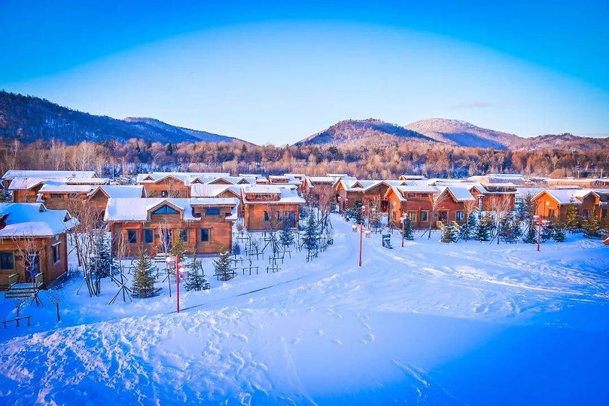 Snow-covered wooden cabins in a winter village, surrounded by trees and mountains under a clear blue sky, evoke a serene, chilly mood.