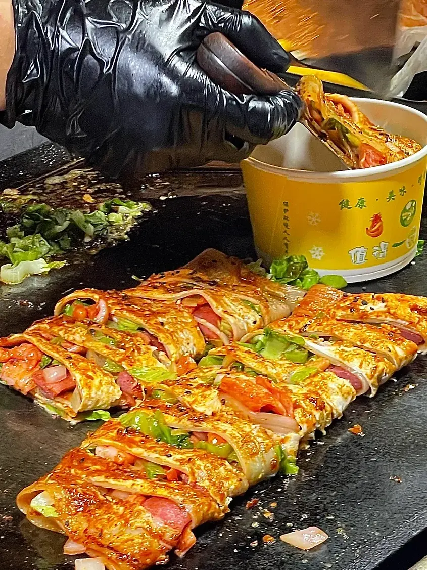 Gloved hand using tongs to serve a sizzling, spicy rolled dish with vegetables and meat into a yellow cup on a grill, with steam rising.
