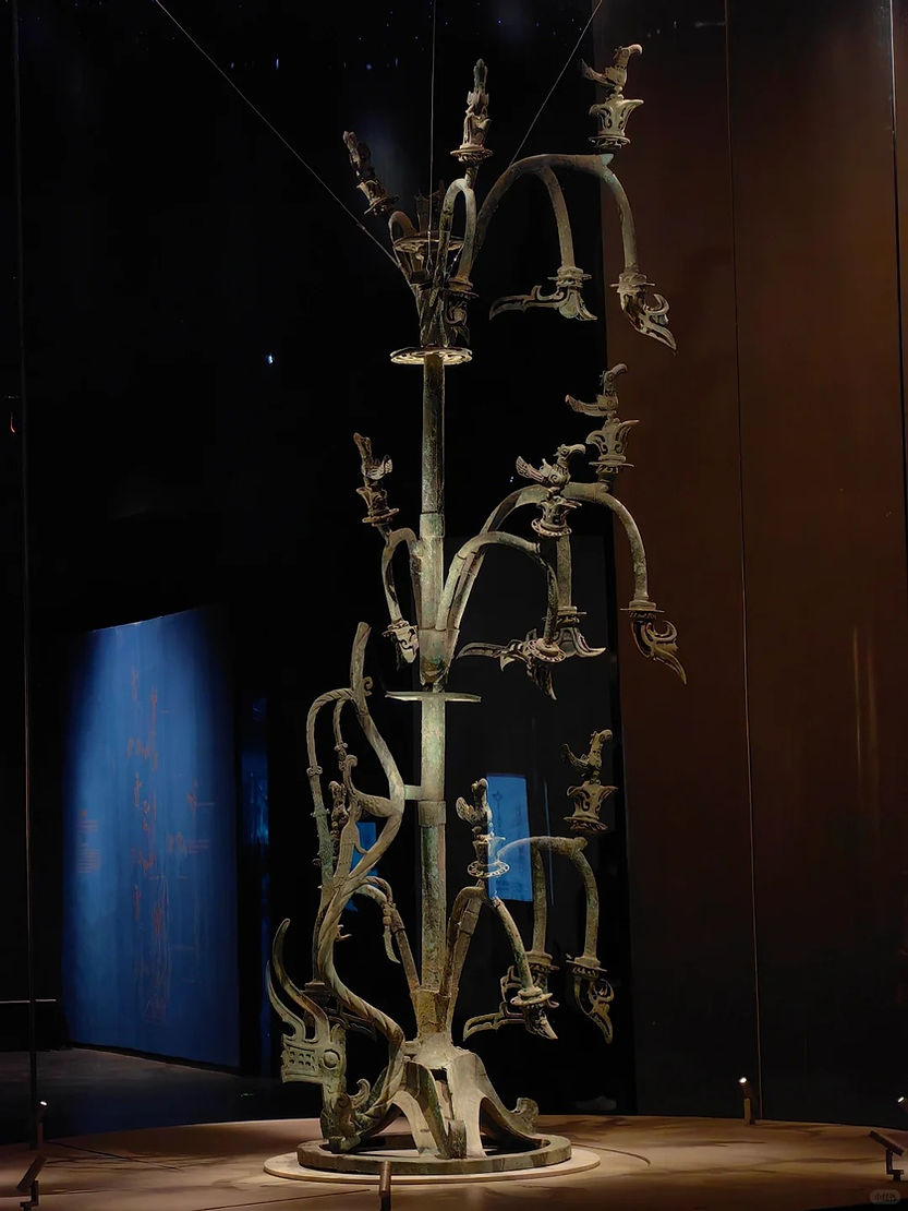 Tall bronze tree sculpture with intricate branches and bird-like figures, set in a dimly lit museum against a dark background.
