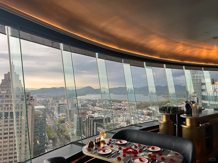 Skyline view from a high-rise with a modern dining setup, rose petals on the table, overlooking a city and distant mountains at sunset.