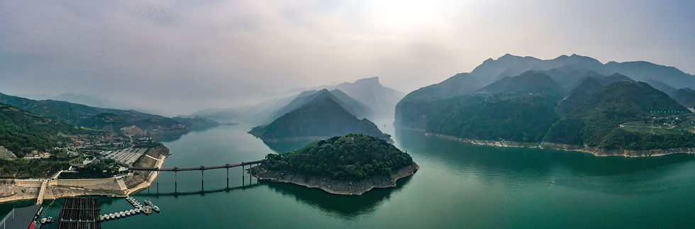 Aerial view of the Three Gorges Dam, China. Green river winds through misty mountains under an overcast sky, featuring docks and a bridge.