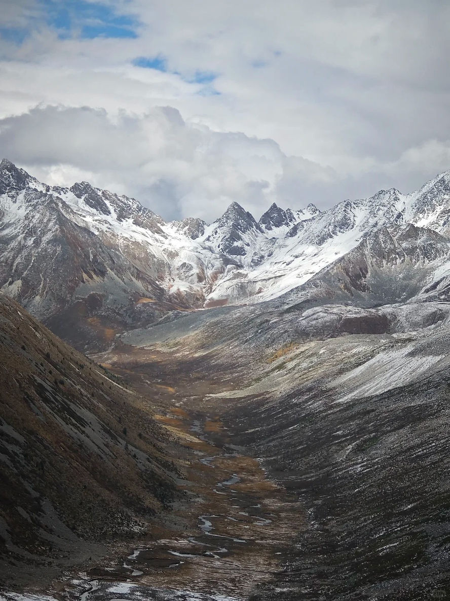 Snow-capped mountains with a cloudy sky, brown valley below with traces of snow, creating a serene and dramatic landscape scene.