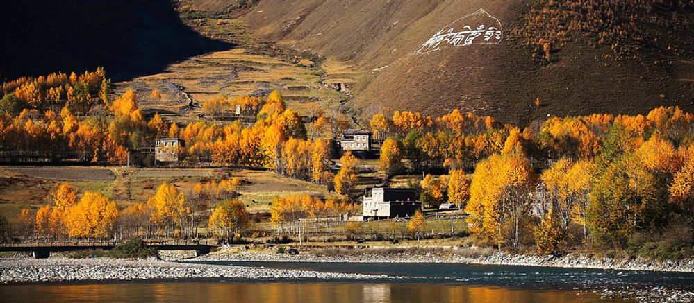 River with autumn trees and small buildings in a valley. Hills with patterns in the background. Warm, peaceful atmosphere.