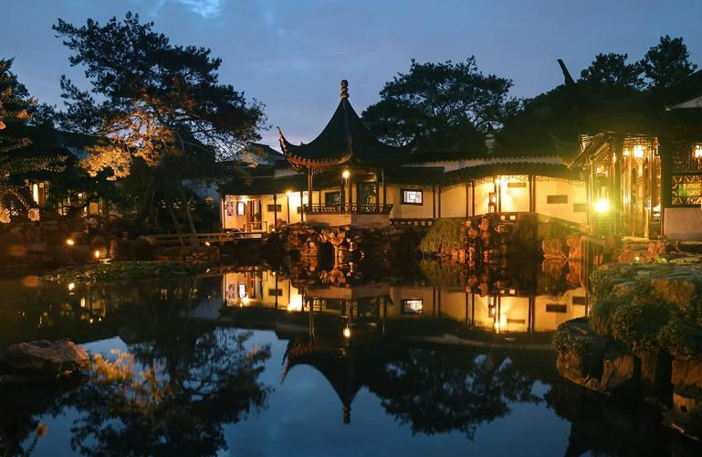 Traditional Chinese garden at dusk with illuminated pavilions and serene pond reflecting trees. Warm lights create a peaceful ambiance.