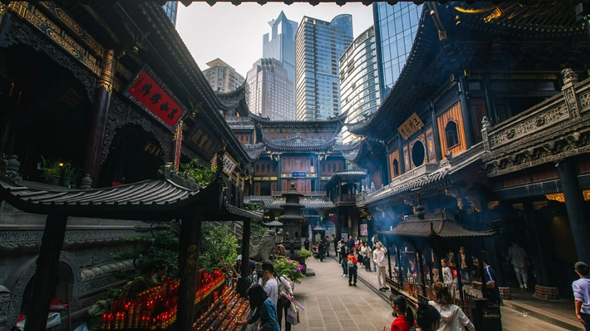 People explore a traditional temple courtyard beneath skyscrapers. Red lanterns and incense create a serene, atmospheric mood.