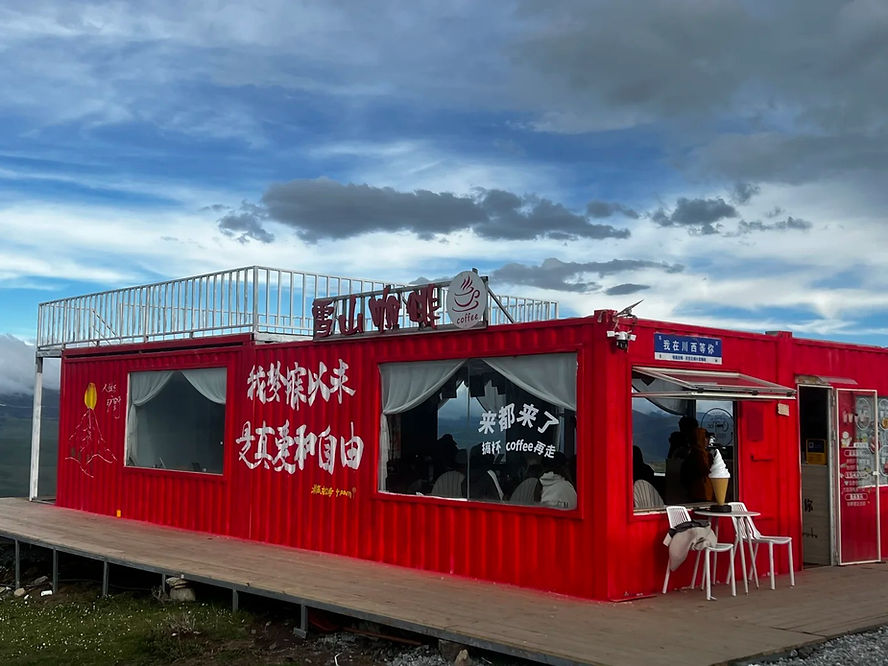 Red shipping container café with white text and mountain design, under a cloudy sky. People inside, chairs outside. Cozy and inviting.