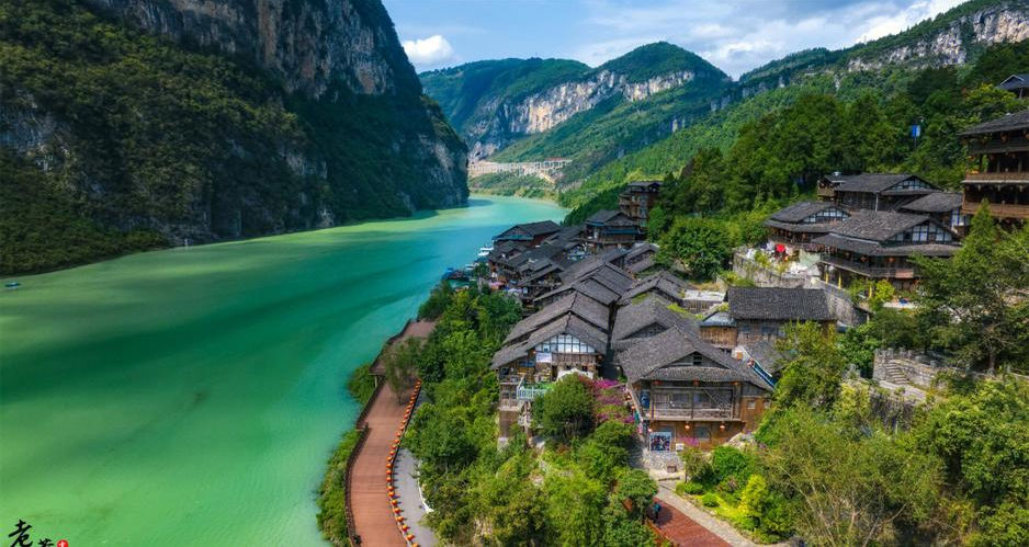 Riverside village with traditional wooden houses along a turquoise river, surrounded by lush green mountains under a blue sky. Serene vibe.