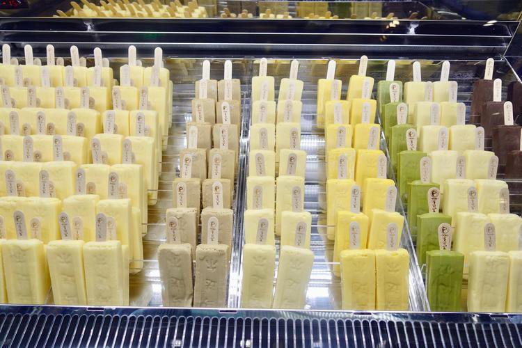 Rows of colorful popsicles in a glass display case, featuring shades of yellow, cream, green, and brown, with visible labels on sticks.