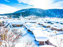 Snow-covered village with wooden houses, red lanterns on trees, and a mountain backdrop. Bright, clear sky creates a serene winter scene.