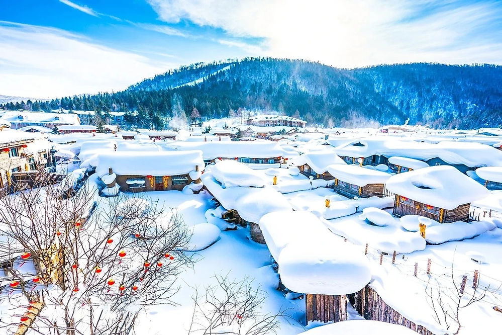 Snow-covered village with wooden houses, red lanterns on trees, and a mountain backdrop. Bright, clear sky creates a serene winter scene.