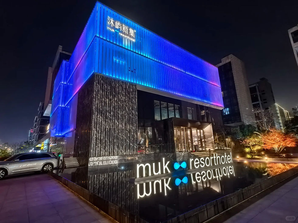 Modern building at night with blue and purple LED lights, glass entrance, and "muk resort hotel" text. Reflective water and parked car.