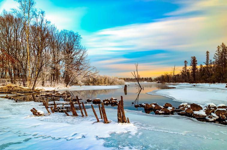 Winter landscape with bare trees by a frozen river reflecting the colorful sunset sky. Snow covers the ground; wooden posts are visible.