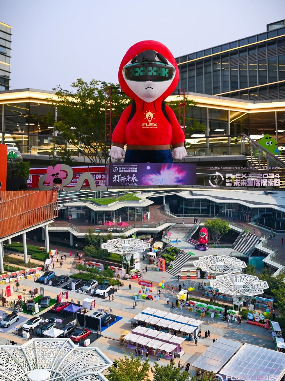 Giant red-hooded figure with VR glasses in outdoor plaza. Surrounding market stalls, colorful booths, trees, and modern buildings. Text: FLEX.