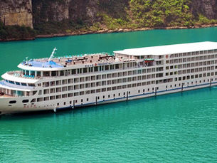 Large white cruise ship sails on turquoise river, flanked by rocky cliffs and lush greenery. Blue accents on ship's deck. Calm mood.