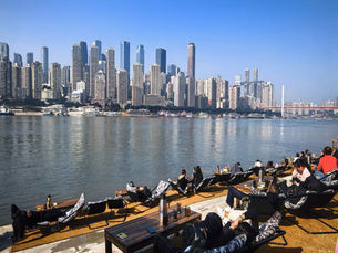 People relax on lounge chairs by a river, with a city skyline in the background. Clear blue sky and calm water set a tranquil mood.