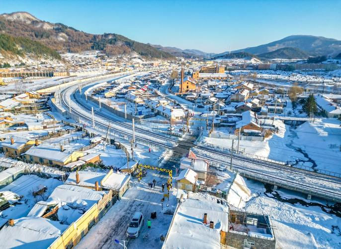 Snowy town with rooftops and streets blanketed in snow, rail tracks curving through. People and cars are in view. Clear blue sky, mountainous backdrop.