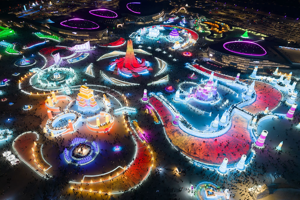 Aerial view of a vibrant ice festival at night with colorful illuminated sculptures, intricate patterns, and crowds of people exploring.