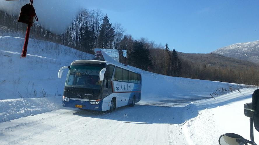 Bus driving on a snowy road with trees and mountains in the background. Clear blue sky, Chinese text on bus side. Winter setting.