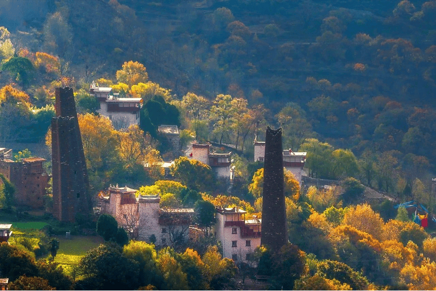 Autumn landscape with tall stone towers among traditional houses, surrounded by vibrant yellow and orange trees. Sunlight creates a warm glow.