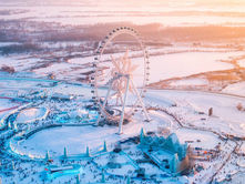Ferris wheel with a snowflake center in a snowy amusement park at sunset. Crowds wander below, surrounded by icy attractions and winter scenery.