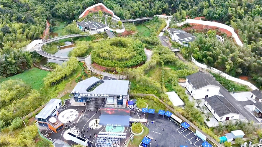 Aerial view of a lush, green panda research center with a circular bamboo garden, modern buildings, and walking paths. Blue umbrellas dot the scene.