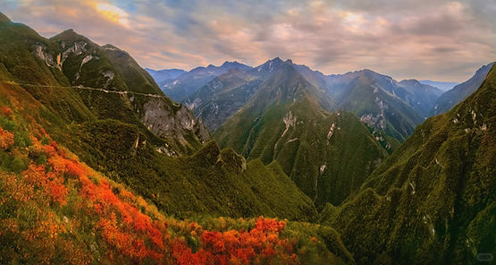 Mountain landscape with lush green hills and vibrant red-orange foliage in the foreground. Soft, cloudy sky creates a serene mood.