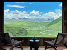 Two chairs and a table on a woven rug face a large window with a view of green hills, snow-capped mountains, and a bright blue sky.