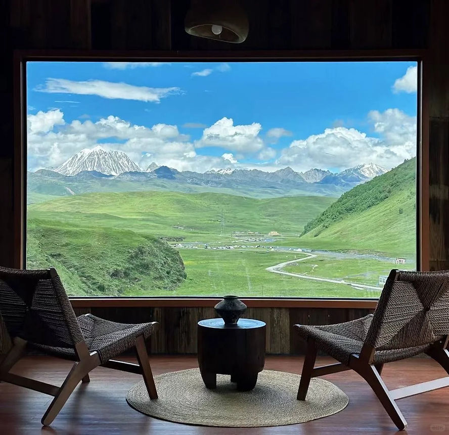 Two chairs and a table on a woven rug face a large window with a view of green hills, snow-capped mountains, and a bright blue sky.