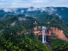 Aerial view of a waterfall cascading down lush green cliffs with misty mountains in the background. The scene is serene and majestic.