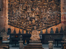 Rows of wooden benches face a seated Buddha statue in a dimly lit temple. A wall filled with small, intricate statues creates a peaceful atmosphere.