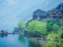 Riverside village with traditional wooden houses, lush green trees, and calm water with small boats docked. Misty mountains in the background.