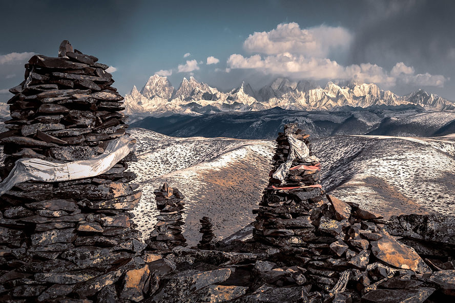 Stone cairns in foreground with snow-dusted hills and distant snowy mountains under a cloudy sky, creating a serene and majestic scene.