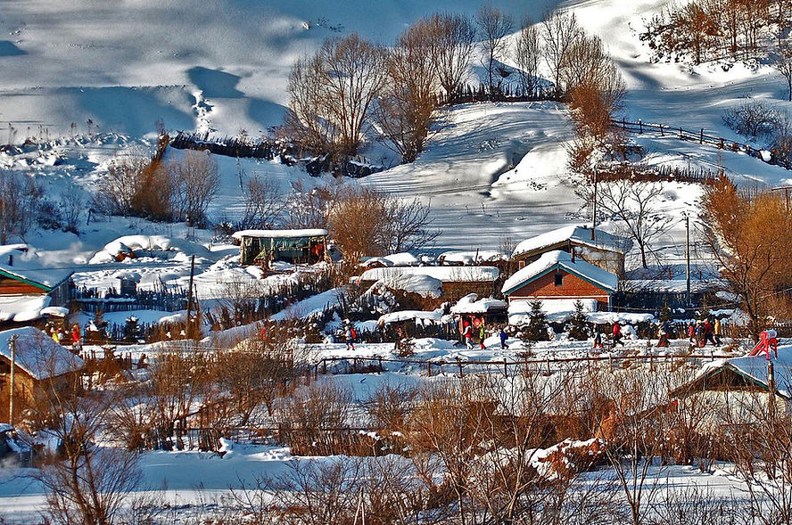 Snowy village landscape with people walking along snow-covered path. Colorful clothing contrasts with white snow, trees scattered around.
