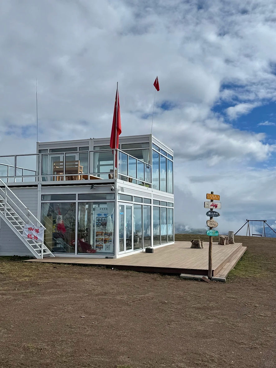 Modern glass building with red flags, set on a grassy hill under a cloudy sky. Signpost points in various directions. Calm atmosphere.
