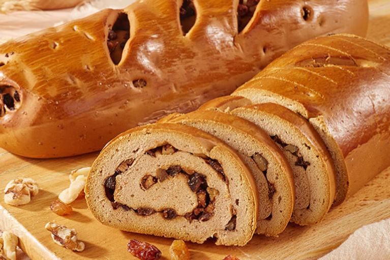 Sliced loaf of brown bread with nuts and raisins, on a wooden board. A whole loaf is in the background. Warm, inviting atmosphere.
