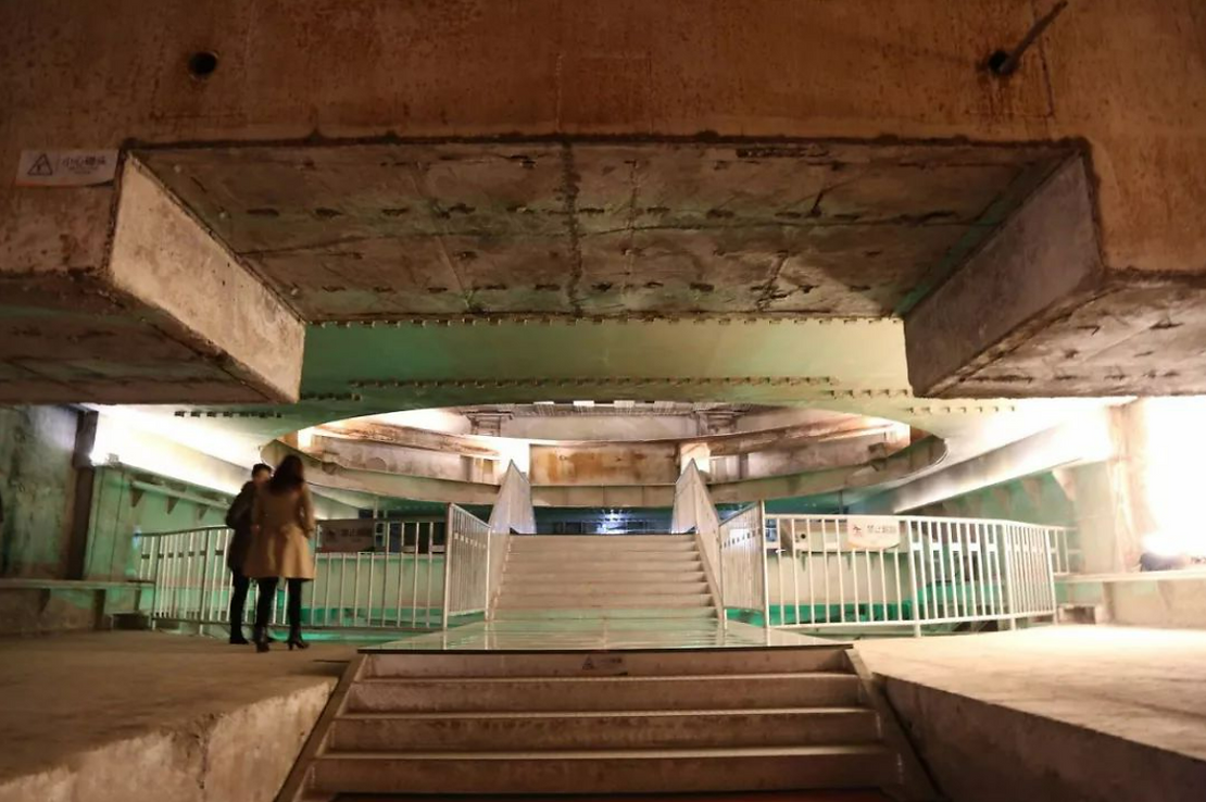 Two people stand near a staircase in a dimly lit, industrial setting with concrete walls and railings. A sign with text is visible above.