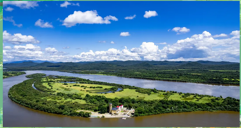 Aerial view of a lush green peninsula surrounded by a winding river under a bright blue sky with fluffy clouds. A small building is visible.