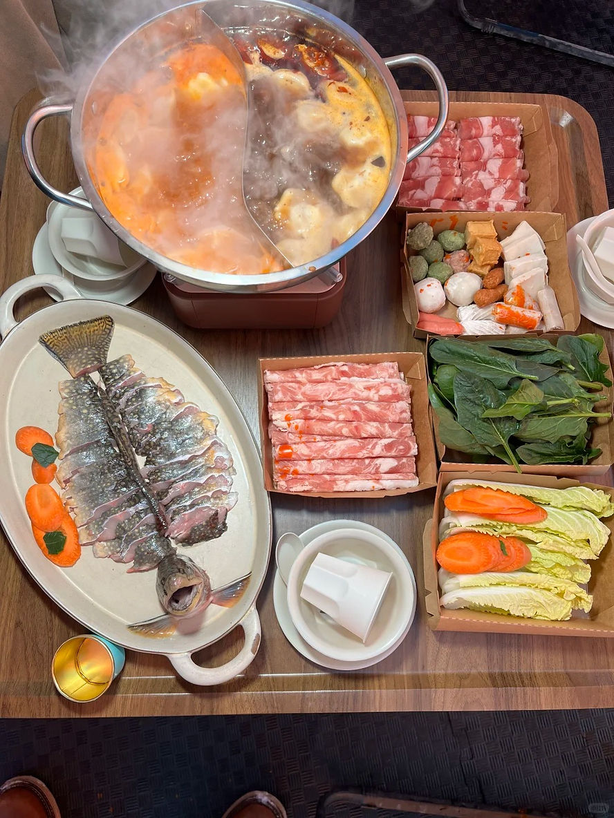 Steaming hot pot with red broth on a wooden table, surrounded by sliced meat, raw fish, vegetables, and dipping sauces. Cozy dining setup.