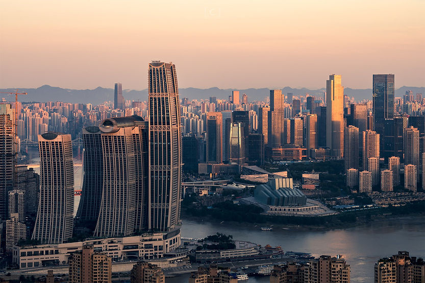 Sunlit skyscrapers in a city skyline at sunset, with a river in the foreground and mountains in the distance. Orange and pink hues dominate.