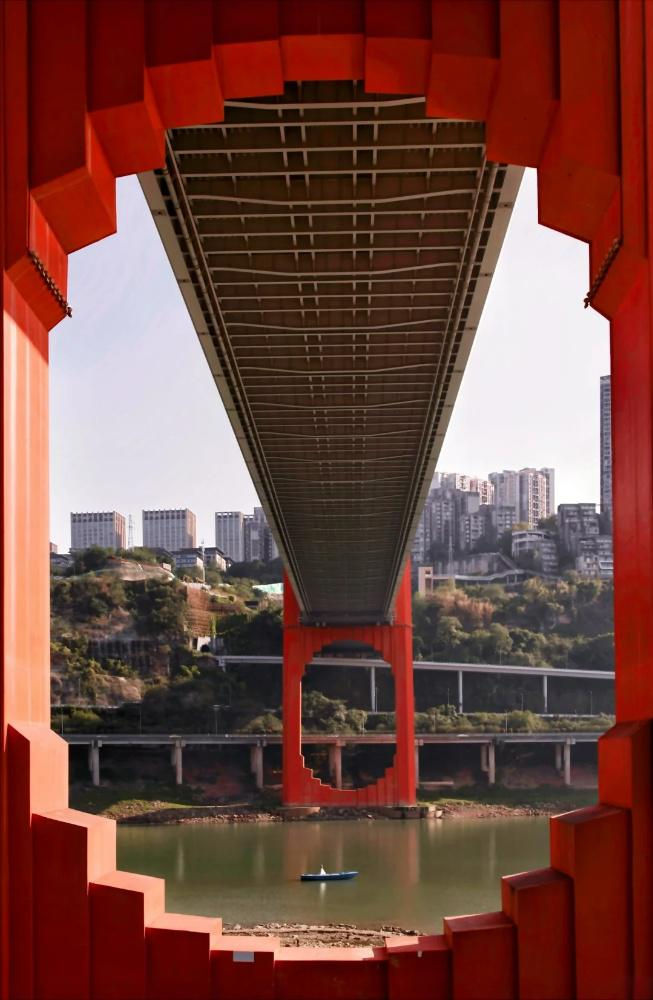 View under a red bridge with a river below and a small boat. City skyline and greenery in the background, framed by bridge architecture.