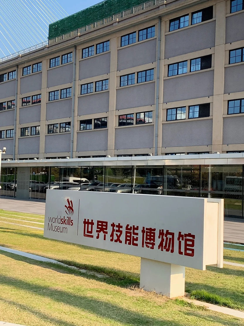 Sign for WorldSkills Museum in front of a large building with rows of windows. Text in English and Chinese. Sunny day, green grass.
