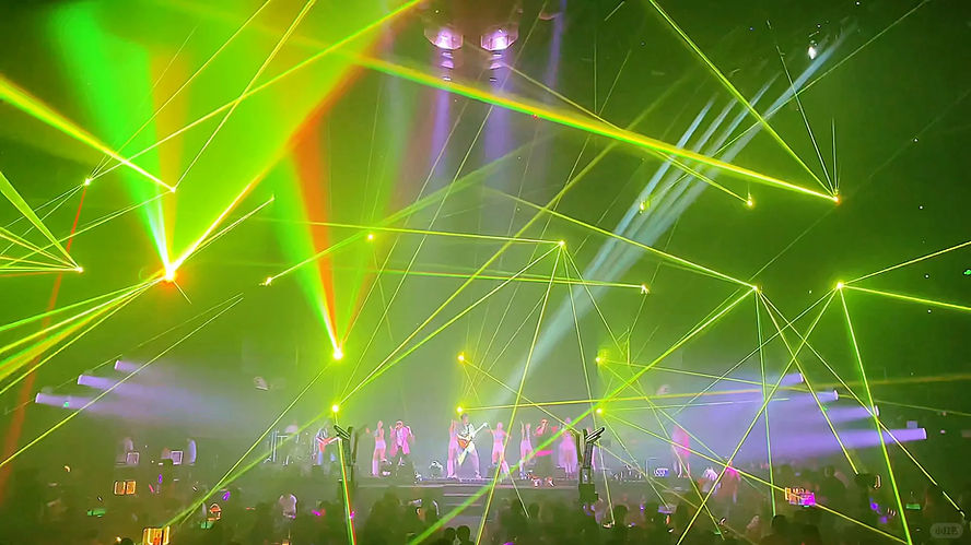 Concert stage with performers, green and yellow laser lights, lively crowd. Energetic atmosphere, no visible text.