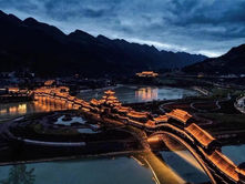 Illuminated traditional bridge at dusk over a calm river, set against silhouetted mountains under a cloudy sky. Warm lights reflect on water.