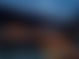 Illuminated traditional bridge at dusk over a calm river, set against silhouetted mountains under a cloudy sky. Warm lights reflect on water.