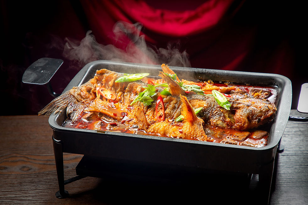 Steaming grilled fish with peppers and herbs in a black pan on a wooden table. Red background adds warmth and richness to the scene.