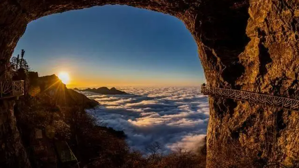 Sunrise over a sea of clouds viewed from a cliffside cave. Golden light, rugged textures, and a bridge enhance the serene atmosphere.